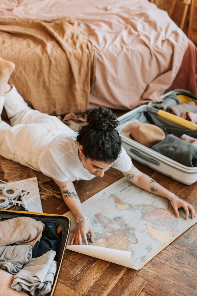 A woman lying on the floor indoors, planning a trip with a map and open suitcase.